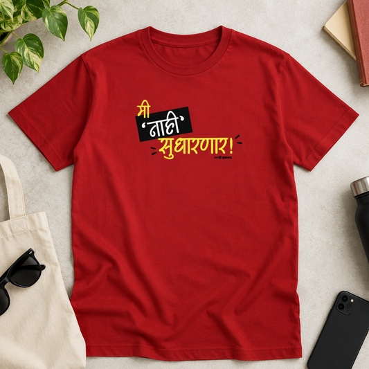 A red short-sleeve t-shirt with Hindi text printed on the chest, styled in a flat-lay arrangement with a canvas tote bag, sunglasses, a black water bottle, a smartphone, and a green plant on a light gray surface.