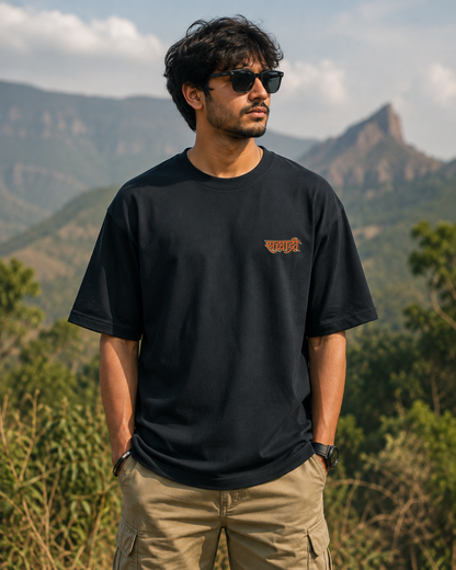 A man wearing a black crewneck t-shirt with a small orange graphic on the left chest, paired with khaki cargo pants and sunglasses, standing outdoors with a mountain landscape in the background.

