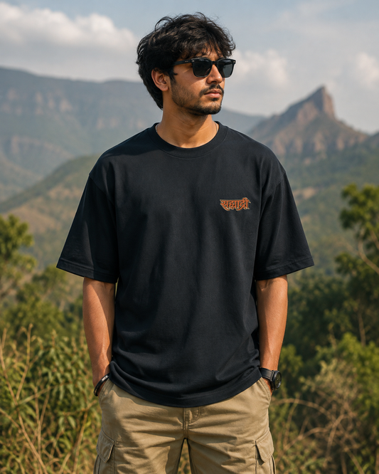 A man wearing a black crewneck t-shirt with a small orange graphic on the left chest, paired with khaki cargo pants and sunglasses, standing outdoors with a mountain landscape in the background.

