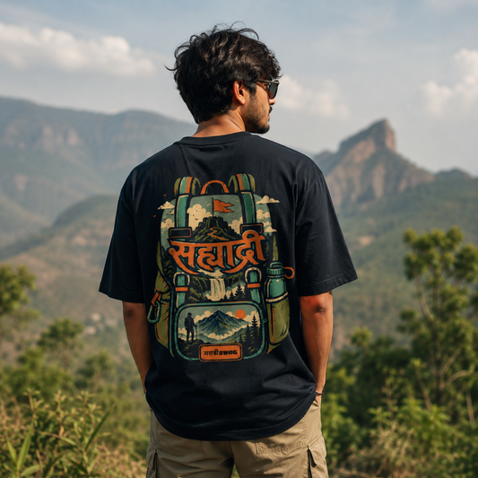 A man stands outdoors in a mountainous landscape, viewed from behind, wearing a black graphic T-shirt with a large illustrated backpack design and Hindi text on the back.
