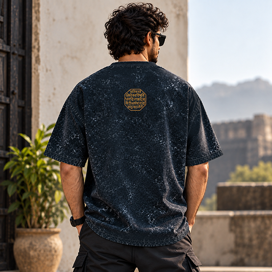 A man viewed from behind wearing a dark navy short-sleeve t-shirt with a subtle speckled texture and a small gold graphic on the upper back. The setting is outdoors with a stone wall, potted plant, and distant mountains in the background.

