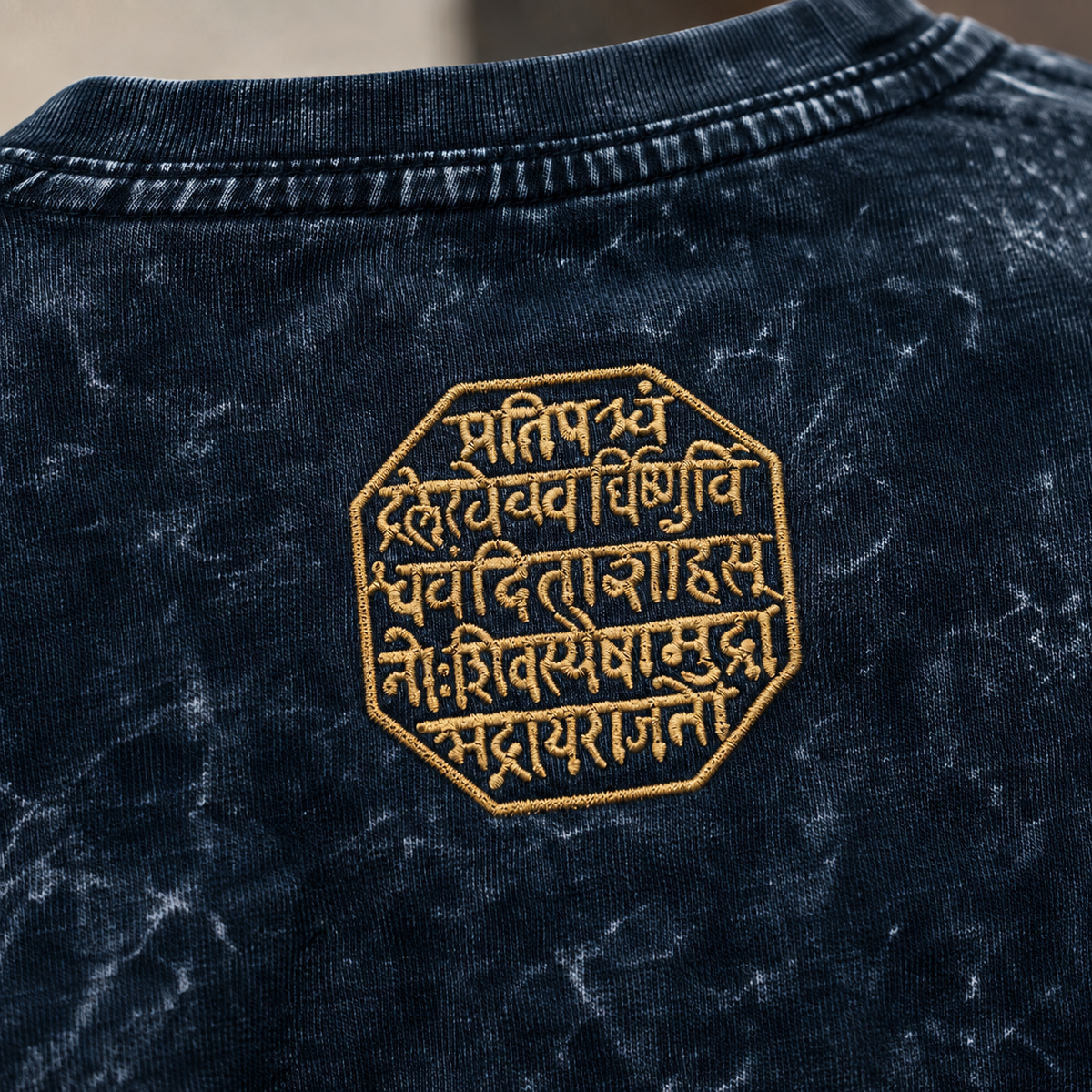 Close-up of the back of a dark navy acid-wash t-shirt featuring a gold embroidered octagonal patch with Sanskrit script.