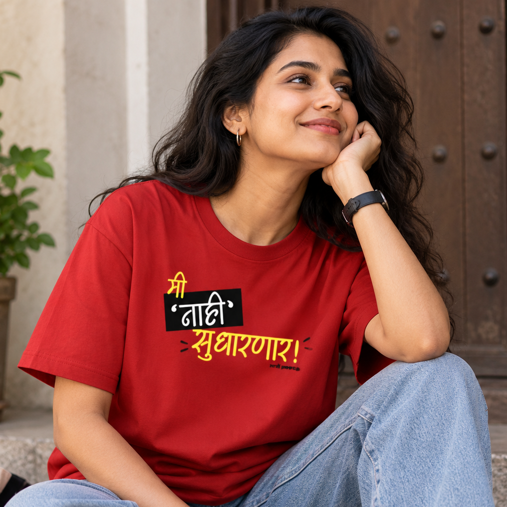 A woman sitting on stone steps wearing a red graphic T-shirt with Marathi text and light blue jeans, posed in front of a wooden door with a potted plant in the background.

